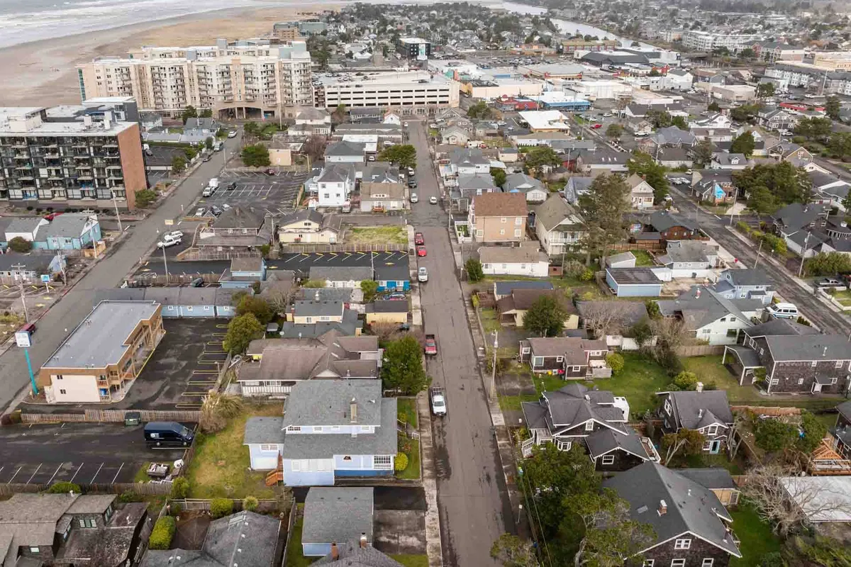 A vibrant neighborhood scene showing both residential homes and commercial buildings, with the ocean visible in the background. The streets are designed for walking, and the area exudes a relaxed, community-oriented vibe. You can see hints of local shops and cafes just beyond the homes, emphasizing the walkable nature of the area.