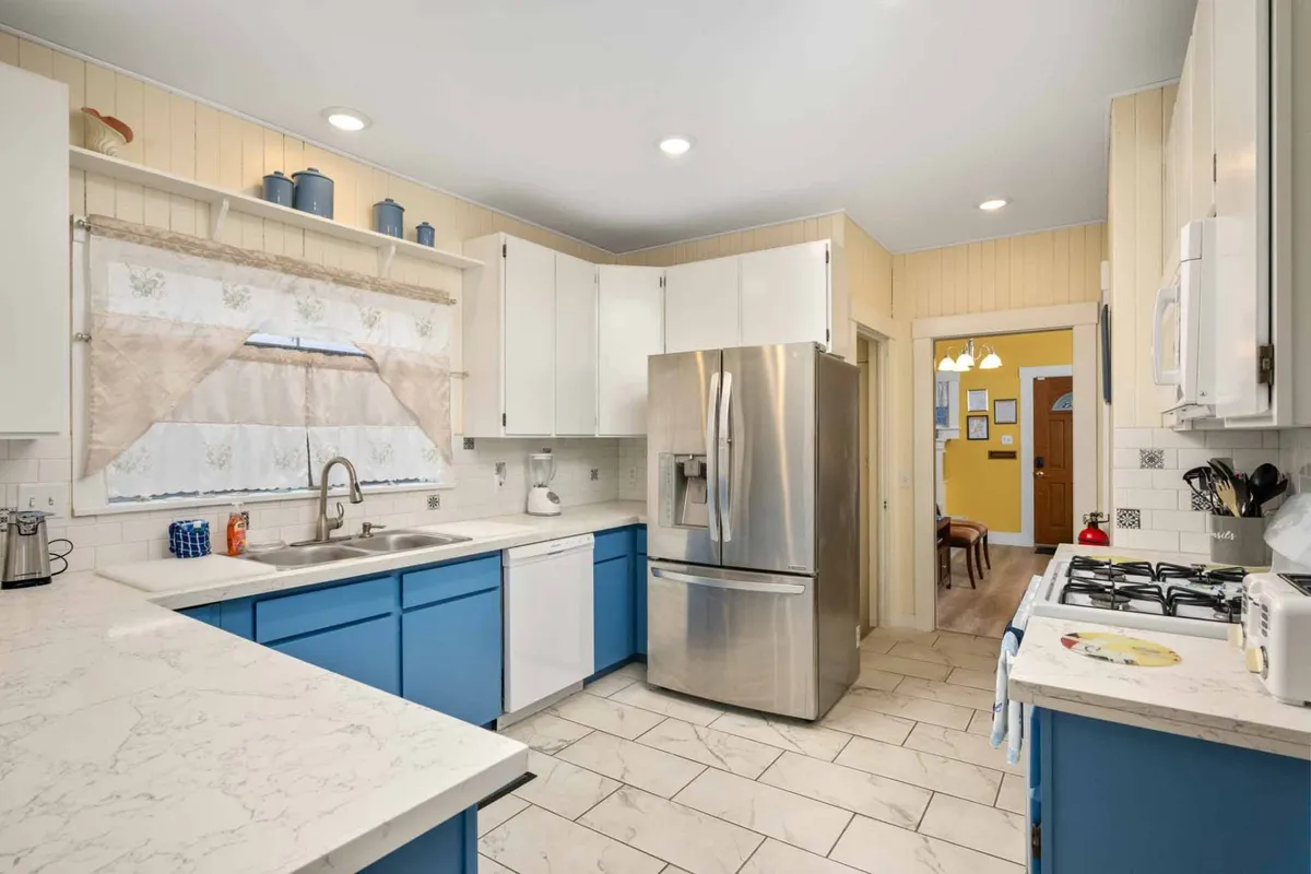 This image captures a bright kitchen featuring blue cabinets, a stainless steel refrigerator, and a marble countertop. The space feels open and inviting, with a layout that encourages socializing and group cooking. Visible kitchen appliances and surfaces suggest readiness for meal prep and family gatherings.