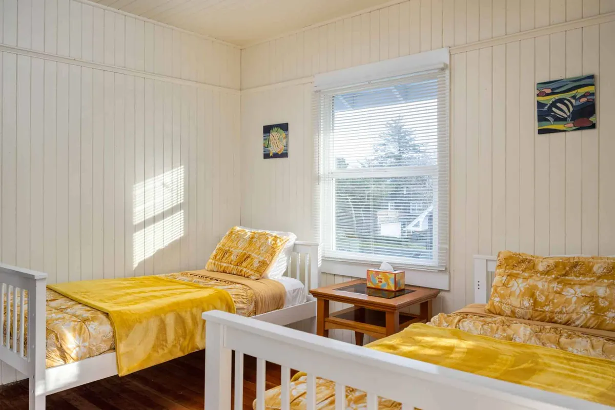 A cheerful bedroom with two beds adorned in bright yellow bedding, a window streaming in sunlight, and a small wooden table with decorative items. The layout is ideal for kids or siblings, offering both comfort and a splash of playful color.