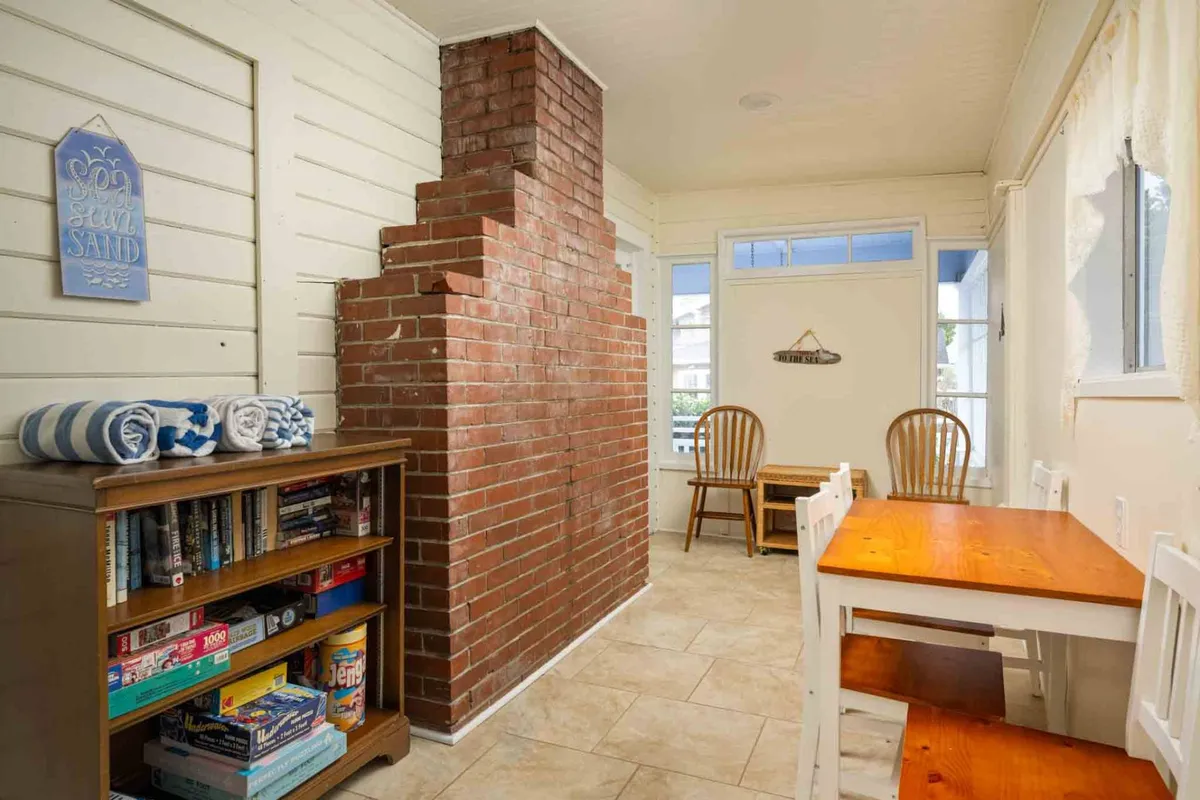 A warm and welcoming interior view, this image centers on a wooden dining table set near a brick chimney and a bookshelf filled with games. The scene exudes comfort and fun, perfect for evenings of poker, puzzles, or sharing meals.