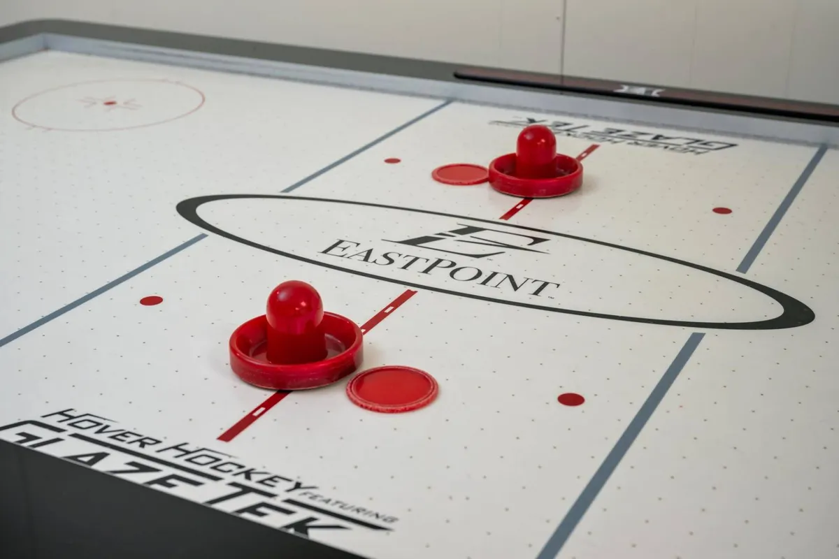Close-up on an energetic hover (air) hockey table with red pucks and a branded logo, set in a well-lit area. The image evokes excitement and draws attention to the active, competitive experiences available in the game room.