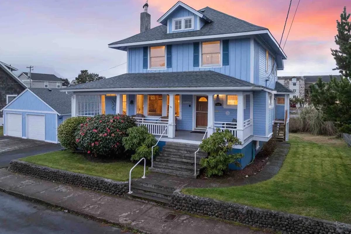 This exterior shot features the blue facade of Roxie's Retreat, a generous front porch perfect for socializing, and a well-maintained garden set against a vibrant sunset sky. The inviting porch hints at a space ideal for gatherings or relaxing after a fun day.