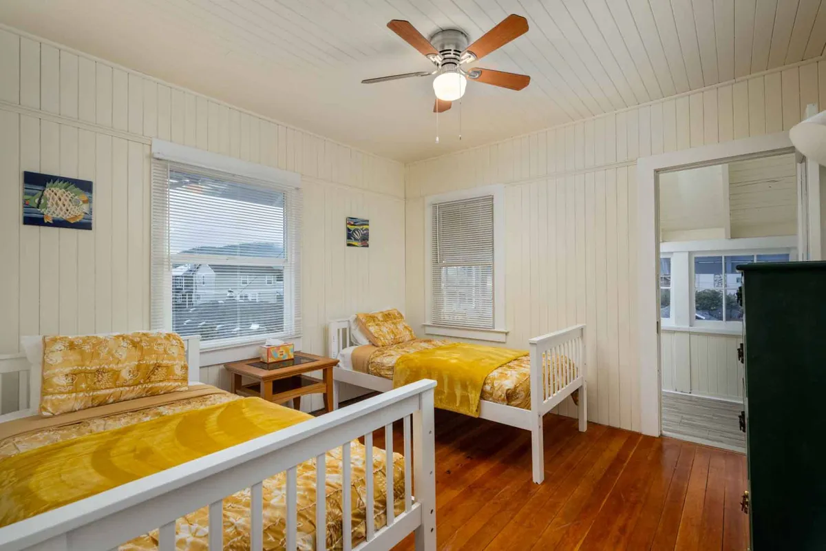 A light-filled bedroom with two single beds featuring yellow bedding, wooden flooring, large windows for ample sunlight, and decorative wall art. A ceiling fan adds extra comfort.