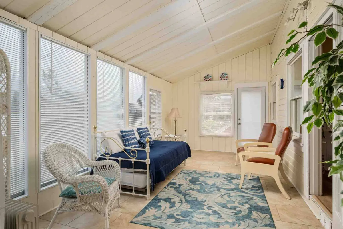 A bright sunroom features a daybed with blue bedding, a woven chair, a potted plant, and large windows letting in natural light. The calm, inviting space is perfect for relaxation, reading, or quiet play, as mentioned in the section.