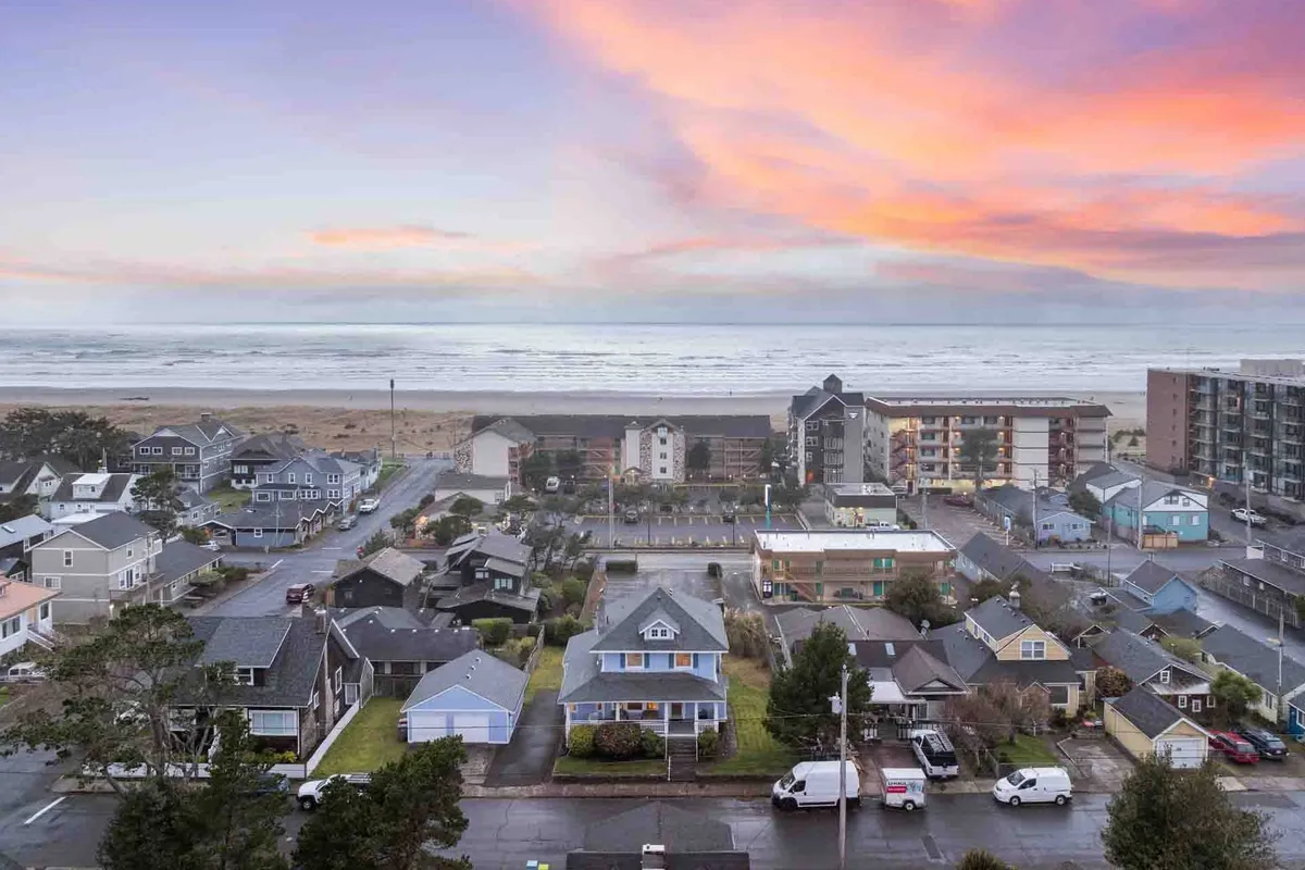 A wide, serene view of the sandy beach stretching alongside the ocean, with gentle waves and colorful sunset sky overhead. Several houses are visible in the background, suggesting the proximity of the neighborhood to the beach. The mood is tranquil, inviting families to enjoy evening walks, kite flying, and beach activities.