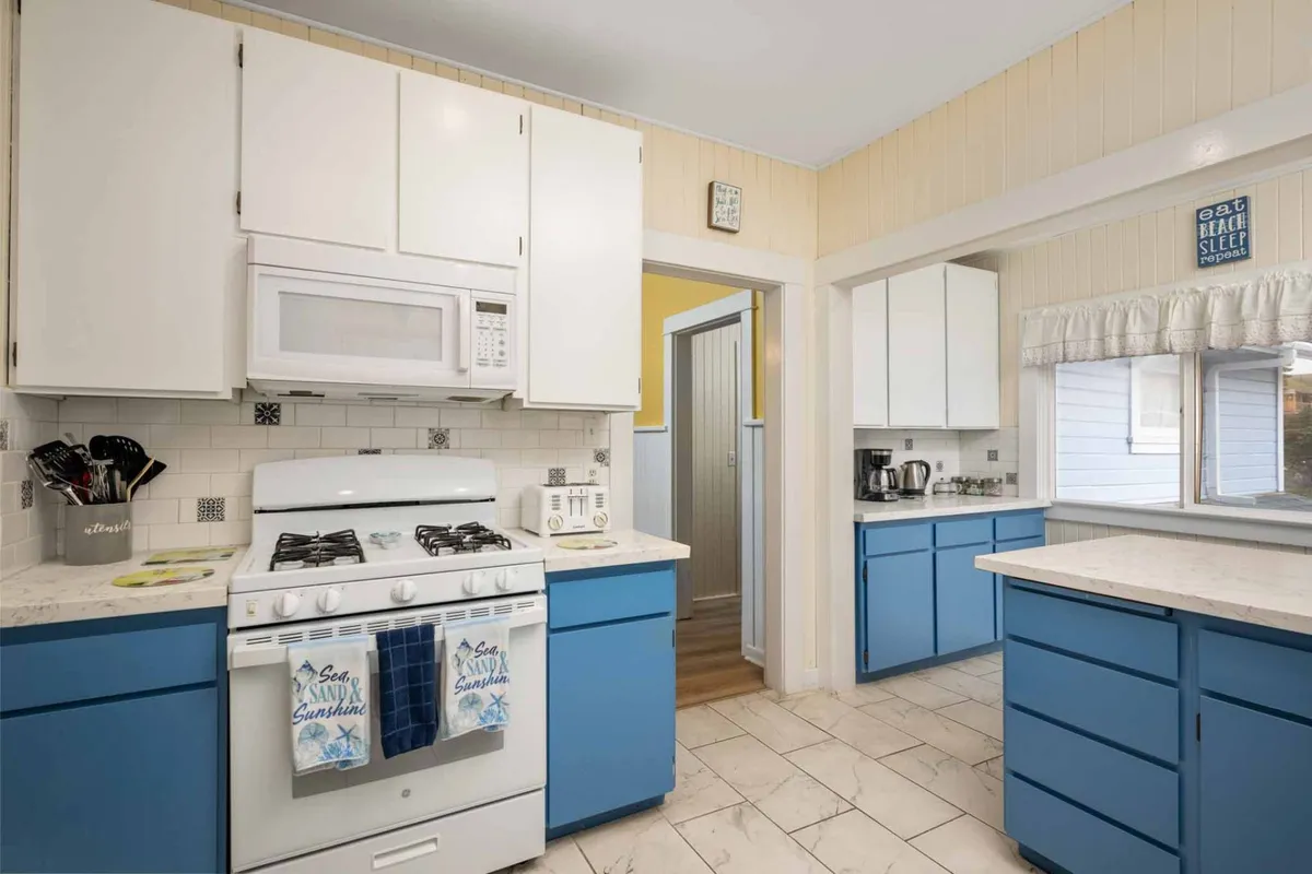 A cozy corner of the kitchen featuring a white gas stove, blue cabinetry, and a sunlit window. Natural light pours in, creating an inviting environment for morning coffee or baking sessions. The setup feels both practical and warm, ideal for daily rituals and spontaneous cooking projects.