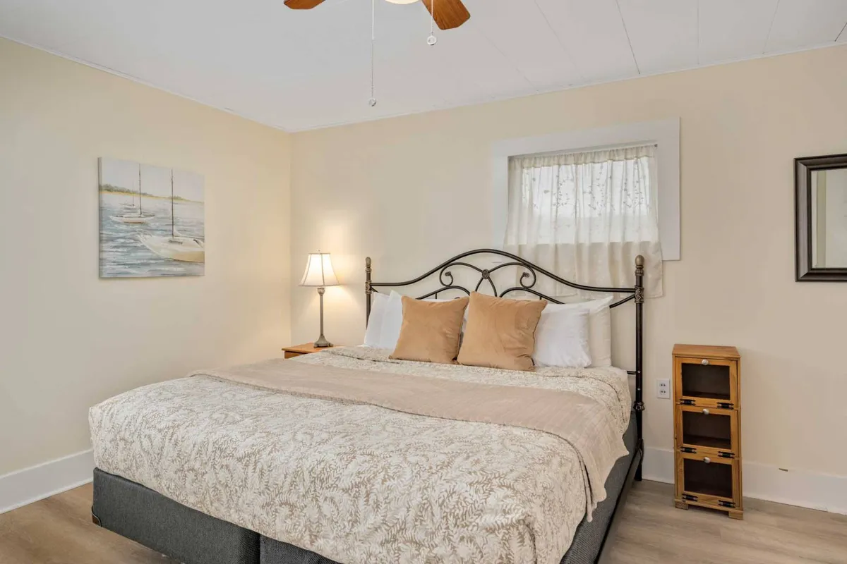 A cozy bedroom with a comfortable bed, soft lamp lighting, a framed painting on the wall, and a window that lets in natural light. The bedding looks plush and inviting, and the room is arranged for maximum comfort and privacy.