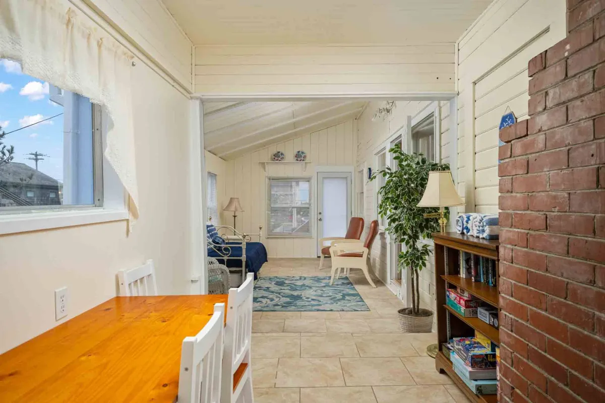 The interior scene showcases a wooden dining table surrounded by comfortable seating in a cozy area, accented by a blue rug and a striking brick wall. Sunlight pours in through nearby windows, inviting guests to linger over board games or stories.